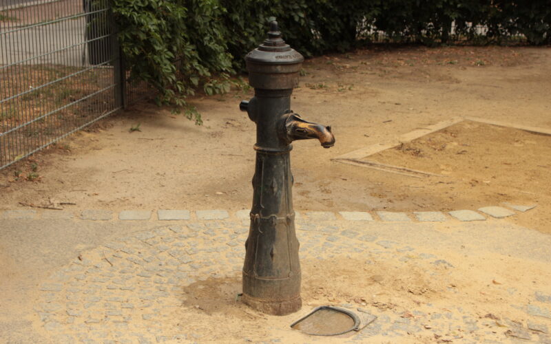 Eine alte Wasserpumpe steht auf einer runden Steinfläche, umgeben von Erde, mit Büschen und einem Metallzaun im Hintergrund.