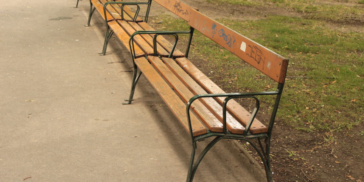 Eine Reihe leerer Holzbänke mit Metallrahmen und einigen Graffiti auf den Rückenlehnen säumen einen gepflasterten Weg in einem Park mit Gras und einem Baum in der Nähe.