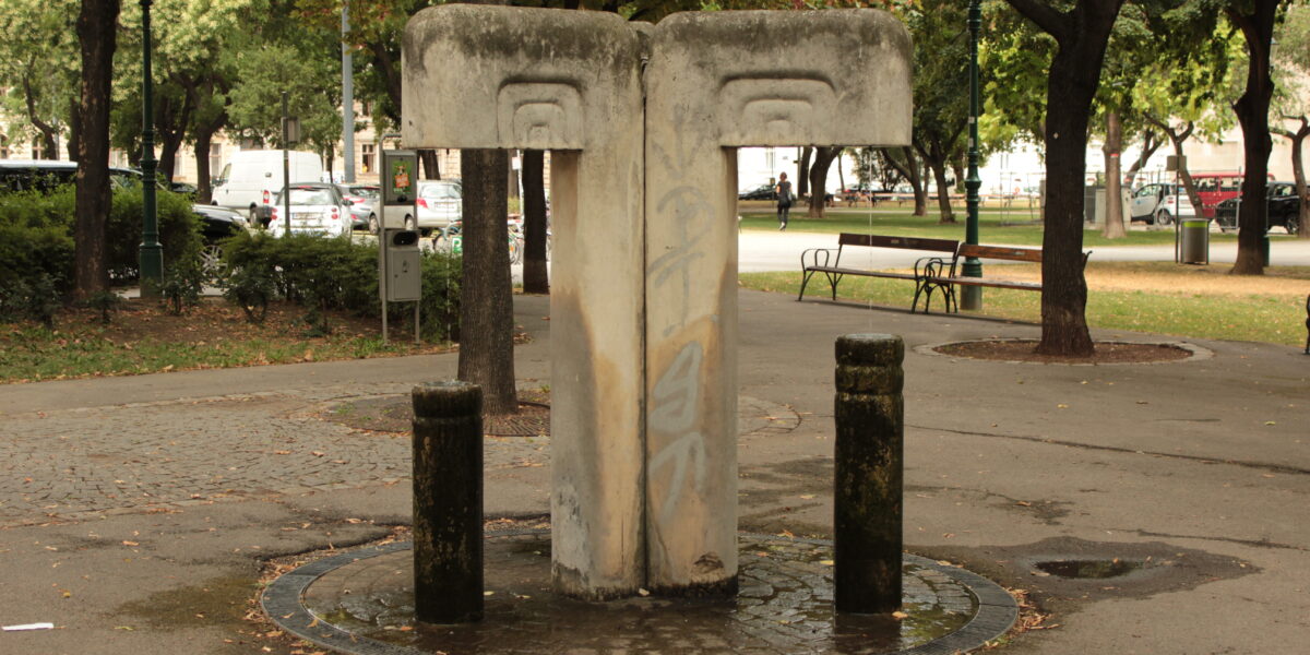 Betontrinkbrunnen mit zwei Ausgüssen und Graffiti, umgeben von Bäumen und Bänken in einem Stadtpark.