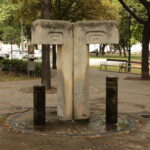 Betontrinkbrunnen mit zwei Ausgüssen und Graffiti, umgeben von Bäumen und Bänken in einem Stadtpark.