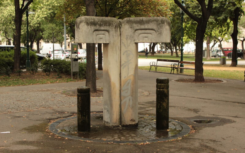 Betontrinkbrunnen mit zwei Ausgüssen und Graffiti, umgeben von Bäumen und Bänken in einem Stadtpark.