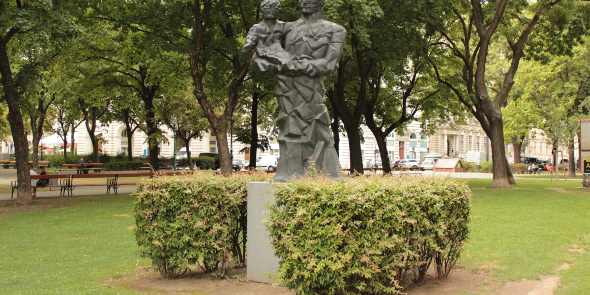 Eine Metallstatue eines Erwachsenen, der ein Kind hält, steht auf einem Sockel, umgeben von Büschen in einem Park mit Bäumen und Bänken.