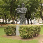 Eine Metallstatue eines Erwachsenen, der ein Kind hält, steht auf einem Sockel, umgeben von Büschen in einem Park mit Bäumen und Bänken.