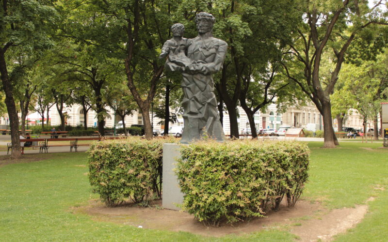 Eine Metallstatue eines Erwachsenen, der ein Kind hält, steht auf einem Sockel, umgeben von Büschen in einem Park mit Bäumen und Bänken.