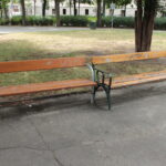 Zwei hölzerne Parkbänke, eine intakt und eine beschädigt, bei der Latten fehlen, stehen nebeneinander auf einer gepflasterten Fläche, umgeben von Gras und Bäumen.