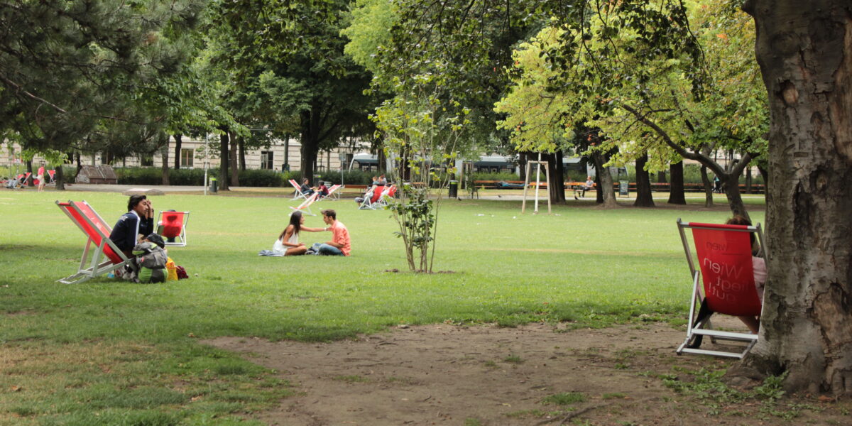 Menschen sitzen und entspannen auf Liegestühlen und Gras in einem grünen Stadtpark mit vereinzelten Bäumen und offenen Rasenflächen.