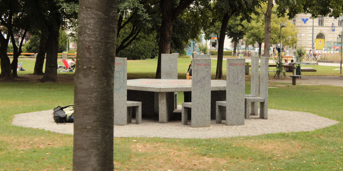 Ein runder Steintisch mit sechs passenden Stühlen steht auf einer kreisförmigen, gepflasterten Fläche in einem Park, umgeben von Gras und Bäumen. Ein Baumstamm verdeckt teilweise die linke Bildseite.