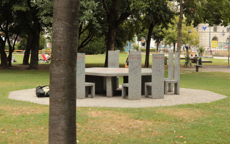 Ein runder Steintisch mit sechs passenden Stühlen steht auf einer kreisförmigen, gepflasterten Fläche in einem Park, umgeben von Gras und Bäumen. Ein Baumstamm verdeckt teilweise die linke Bildseite.