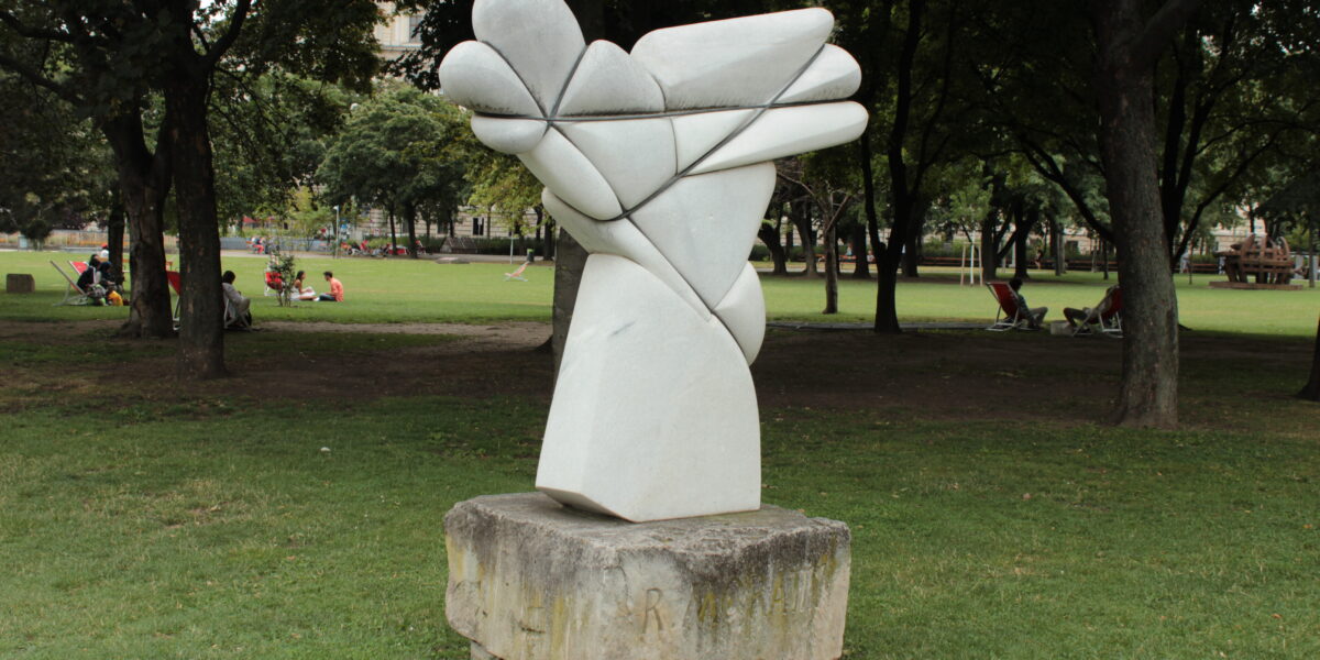 Abstrakte Steinskulptur mit geschwungenen, sich kreuzenden Formen auf einem Betonsockel, aufgestellt auf einer Rasenfläche in einem Park mit Bäumen und Menschen im Hintergrund.