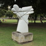 Abstrakte Steinskulptur mit geschwungenen, sich kreuzenden Formen auf einem Betonsockel, aufgestellt auf einer Rasenfläche in einem Park mit Bäumen und Menschen im Hintergrund.