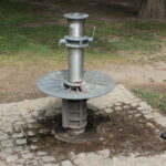 Auf einem Kopfsteinpflaster mit nassen Stellen steht eine öffentliche Wasserpumpe aus Metall; in der Nähe läuft eine Taube auf dem Gras.