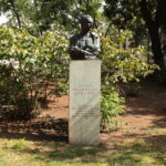 Eine Bronzebüste von Guido Holzknecht auf einem Steinsockel in einem Park mit Bäumen und Büschen im Hintergrund. Auf dem Sockel sind sein Name, sein Lebensjahr und seine Leistungen eingraviert.