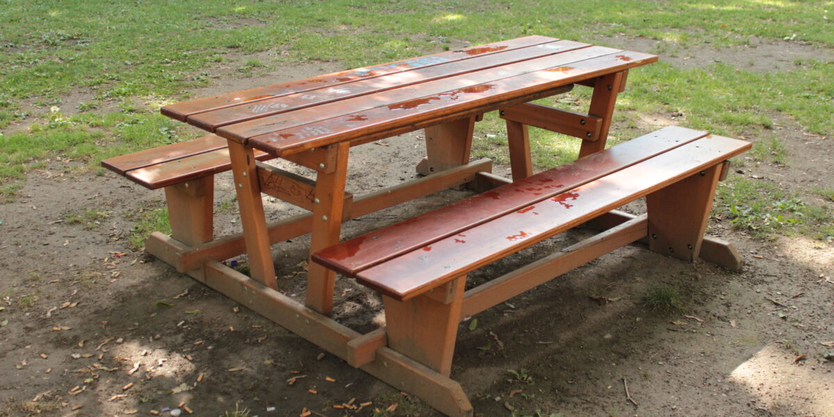 Ein hölzerner Picknicktisch mit daran befestigten Bänken steht auf einem Stück Erde und Gras im Freien.