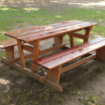 Ein hölzerner Picknicktisch mit daran befestigten Bänken steht auf einem Stück Erde und Gras im Freien.