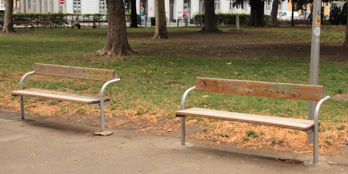Zwei leere Holzbänke mit Metallrahmen stehen auf einem gepflasterten Weg in einem Park, umgeben von Gras, Bäumen und einigen verstreuten Blättern.