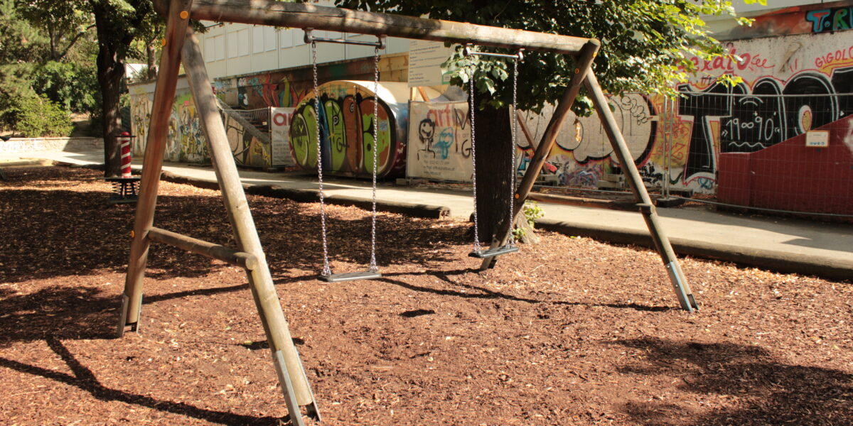 Holzschaukel mit zwei Schaukeln auf einem mit Holzspänen bedeckten Spielplatz, mit Bäumen und einer mit Graffiti bedeckten Mauer im Hintergrund.