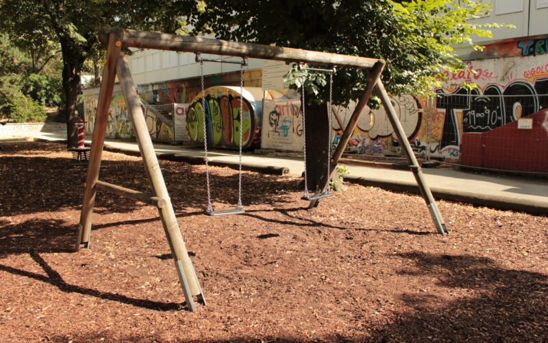 Holzschaukel mit zwei Schaukeln auf einem mit Holzspänen bedeckten Spielplatz, mit Bäumen und einer mit Graffiti bedeckten Mauer im Hintergrund.