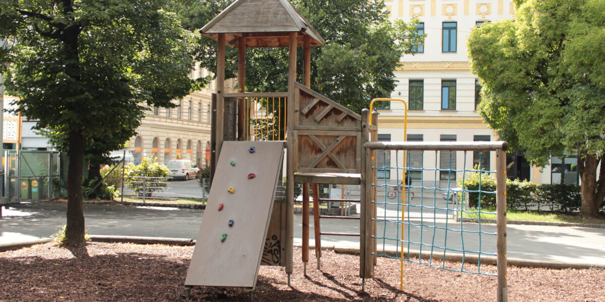 Ein hölzernes Spielgerät mit Rutsche, Kletterwand, Seilnetz und Leiter steht auf Rindenmulch in einem von Bäumen und Gebäuden umgebenen Stadtpark.