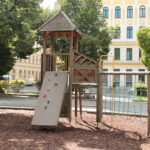 Ein hölzernes Spielgerät mit Rutsche, Kletterwand, Seilnetz und Leiter steht auf Rindenmulch in einem von Bäumen und Gebäuden umgebenen Stadtpark.