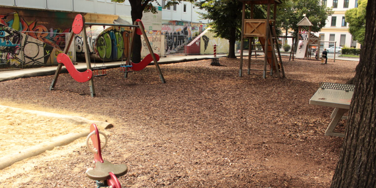 Ein leerer Spielplatz mit Holzspänen als Bodenbelag, verschiedenen Spielgeräten und mit Graffiti beschmierten Wänden im Hintergrund an einem sonnigen Tag.