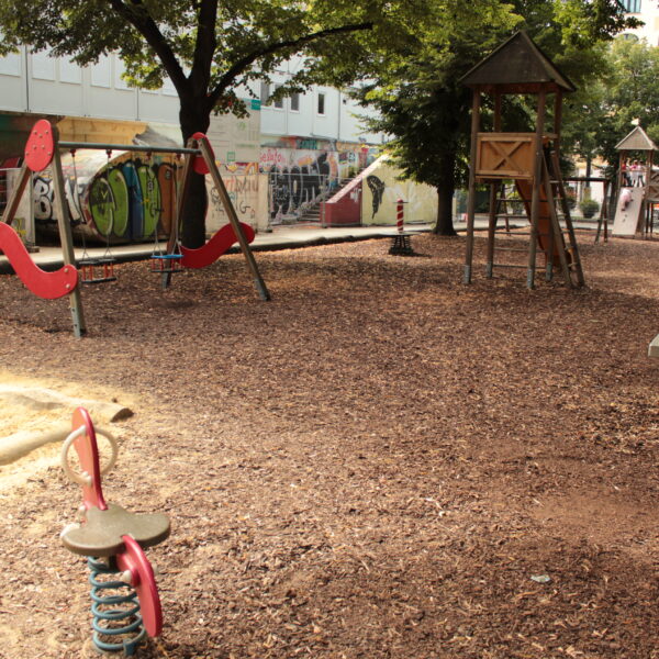 Ein leerer Spielplatz mit Holzspänen als Bodenbelag, verschiedenen Spielgeräten und mit Graffiti beschmierten Wänden im Hintergrund an einem sonnigen Tag.