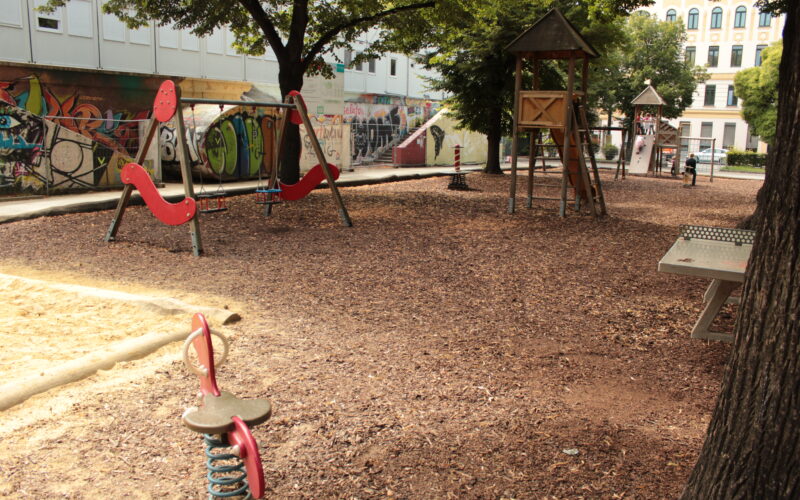Ein leerer Spielplatz mit Holzspänen als Bodenbelag, verschiedenen Spielgeräten und mit Graffiti beschmierten Wänden im Hintergrund an einem sonnigen Tag.