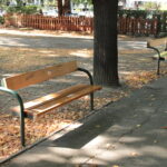Zwei Holzbänke stehen entlang eines gepflasterten Weges in einem Park. Auf dem Boden liegen verstreut Blätter, und Bäume spenden teilweise Schatten.