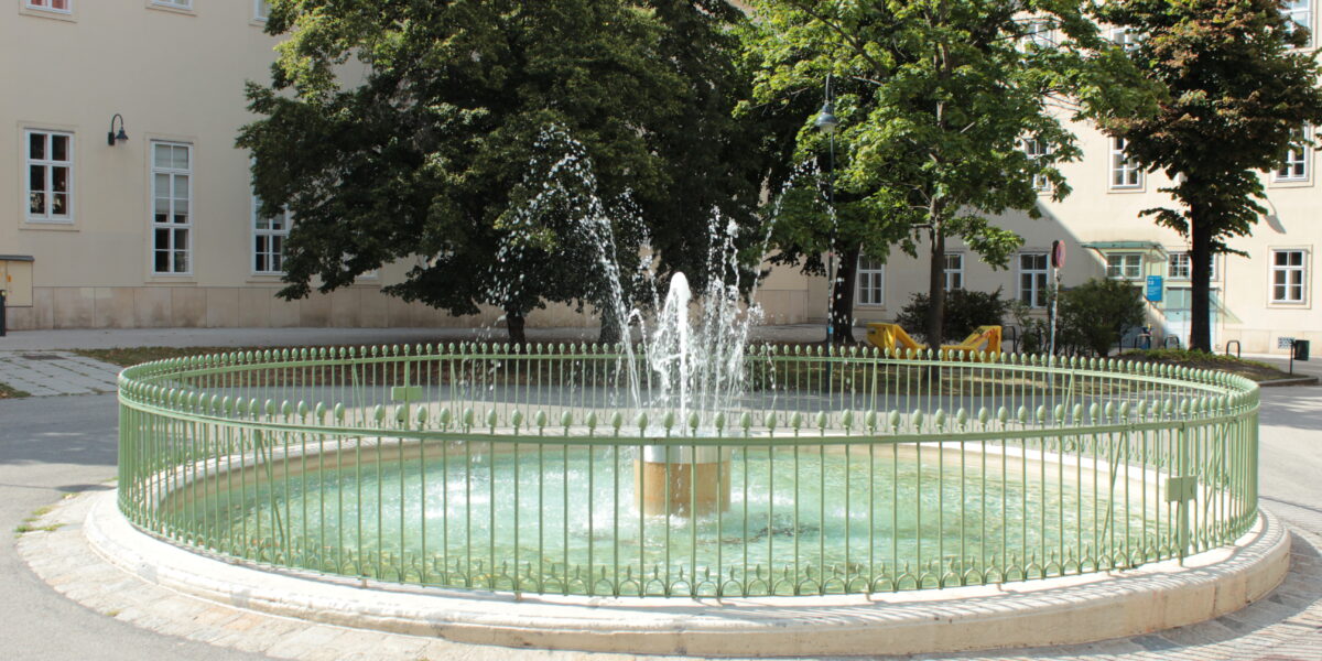 Ein kleiner runder Brunnen mit Wasserstrahlen ist von einem grünen Metallzaun umgeben und befindet sich in einem gepflasterten Innenhof mit Bäumen und Gebäuden im Hintergrund.