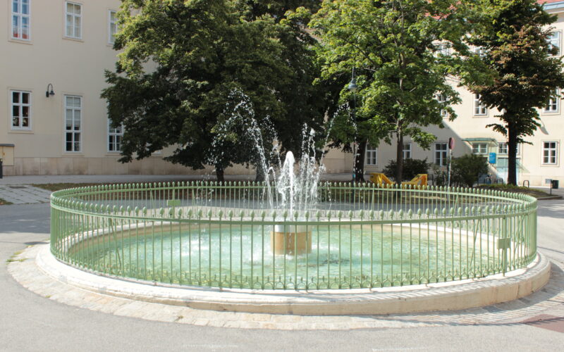 Ein kleiner runder Brunnen mit Wasserstrahlen ist von einem grünen Metallzaun umgeben und befindet sich in einem gepflasterten Innenhof mit Bäumen und Gebäuden im Hintergrund.