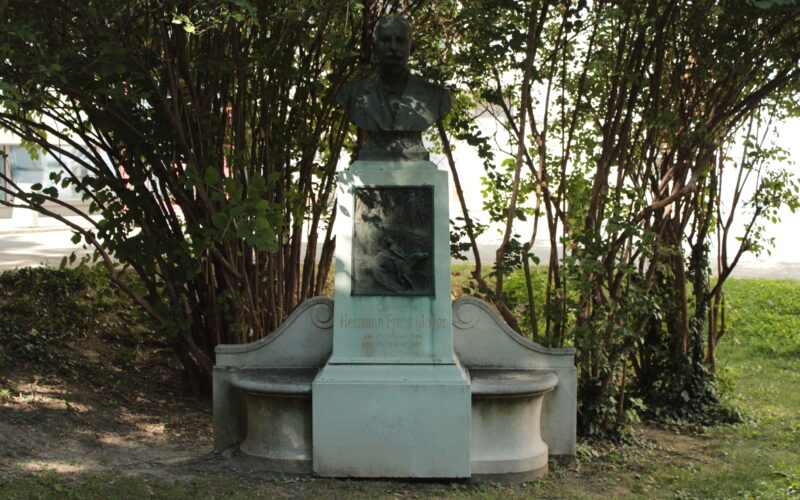 Bronzenes Büstendenkmal mit gravierter Gedenktafel und zwei geschwungenen Bänken, umgeben von Laubbäumen in einer Gartenanlage.