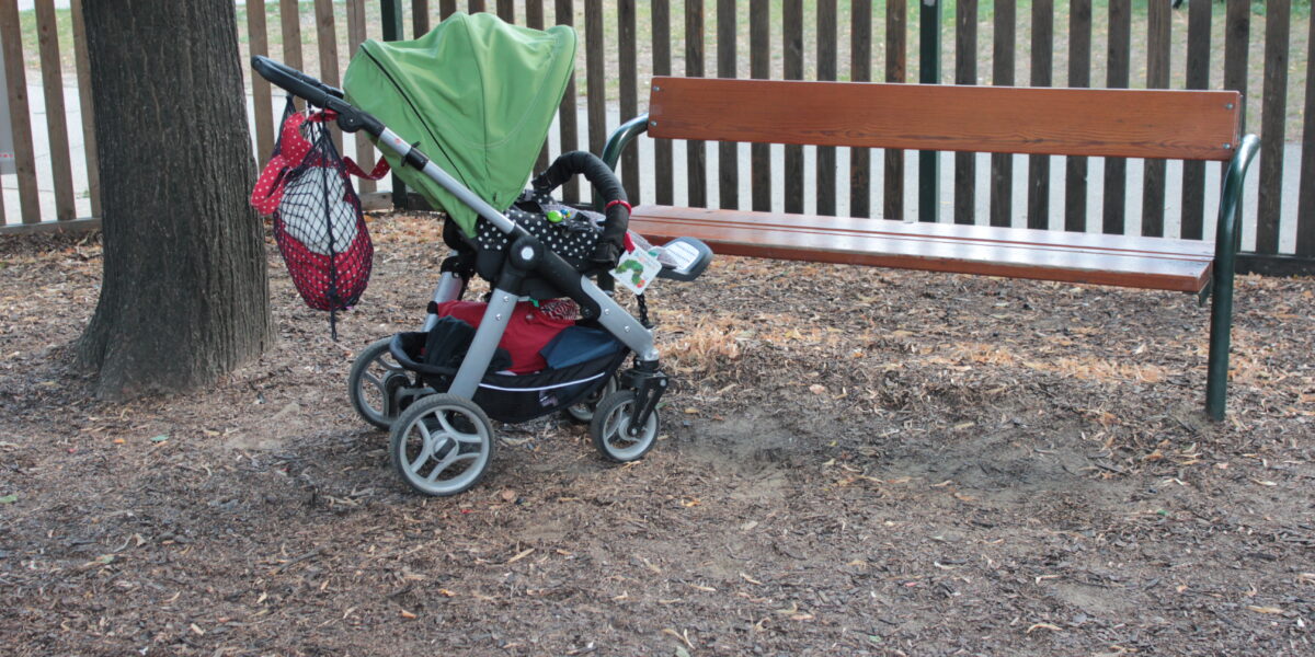 Ein Kinderwagen mit grünem Vordach wird neben einer Holzbank in einem Park abgestellt. Am Griff des Kinderwagens hängt eine Netztasche.
