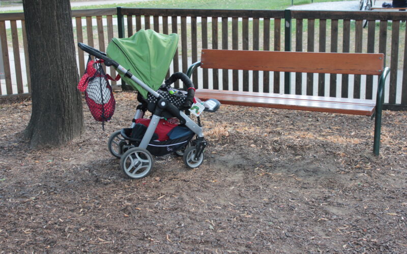 Ein Kinderwagen mit grünem Vordach wird neben einer Holzbank in einem Park abgestellt. Am Griff des Kinderwagens hängt eine Netztasche.