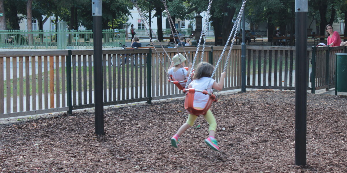 Zwei kleine Kinder sitzen auf einer Schaukel auf einem Spielplatz, umgeben von Holzspänen, mit Erwachsenen und anderen Kindern im Hintergrund.