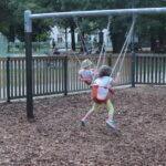 Zwei kleine Kinder sitzen auf einer Schaukel auf einem Spielplatz, umgeben von Holzspänen, mit Erwachsenen und anderen Kindern im Hintergrund.
