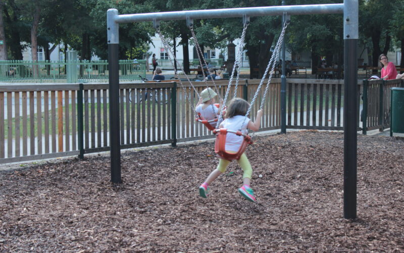 Zwei kleine Kinder sitzen auf einer Schaukel auf einem Spielplatz, umgeben von Holzspänen, mit Erwachsenen und anderen Kindern im Hintergrund.