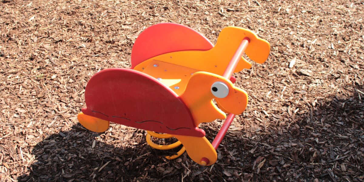 Orangefarbene und rote gefederte Spielplatzwippe in Form eines Dinosauriers auf einem Holzspanboden in hellem Sonnenlicht.