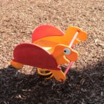 Orangefarbene und rote gefederte Spielplatzwippe in Form eines Dinosauriers auf einem Holzspanboden in hellem Sonnenlicht.