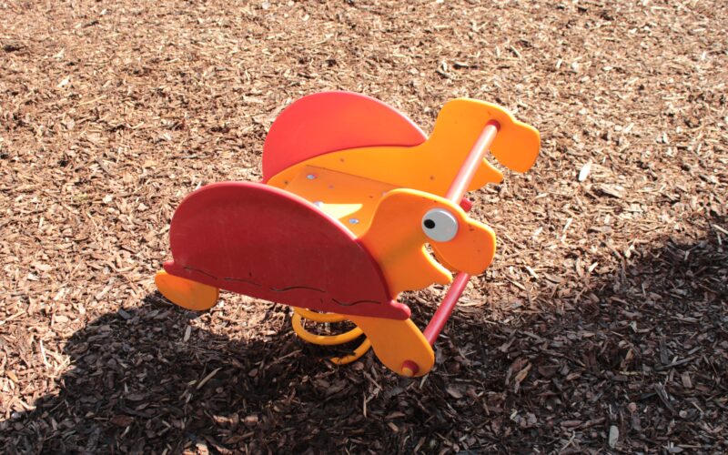 Orangefarbene und rote gefederte Spielplatzwippe in Form eines Dinosauriers auf einem Holzspanboden in hellem Sonnenlicht.