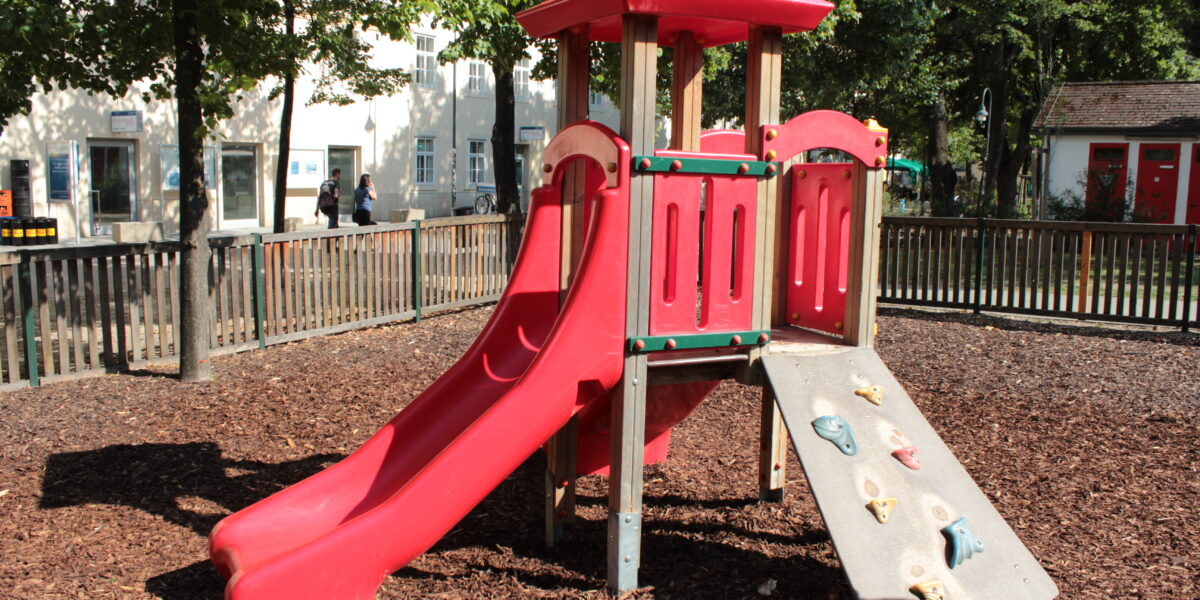 Rote Spielplatzstruktur mit Rutsche und Kletterwand, umgeben von einem Holzzaun und Mulch, mit Bäumen und Gebäuden im Hintergrund.