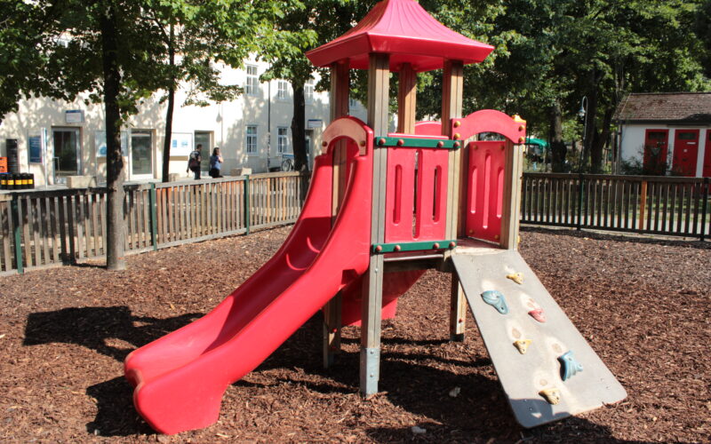 Rote Spielplatzstruktur mit Rutsche und Kletterwand, umgeben von einem Holzzaun und Mulch, mit Bäumen und Gebäuden im Hintergrund.