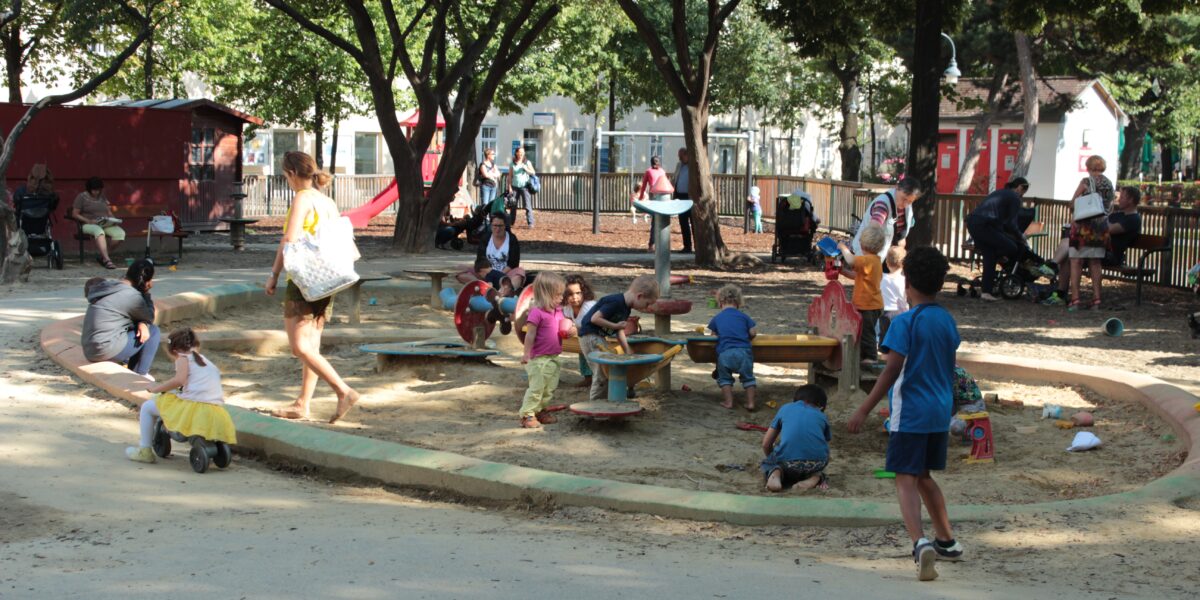 Kinder und Erwachsene befinden sich auf einem Spielplatz mit Sand und Spielgeräten, der an einem sonnigen Tag von Bäumen und Bänken umgeben ist.