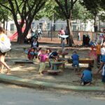 Kinder und Erwachsene befinden sich auf einem Spielplatz mit Sand und Spielgeräten, der an einem sonnigen Tag von Bäumen und Bänken umgeben ist.