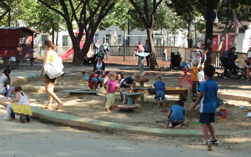 Kinder und Erwachsene befinden sich auf einem Spielplatz mit Sand und Spielgeräten, der an einem sonnigen Tag von Bäumen und Bänken umgeben ist.