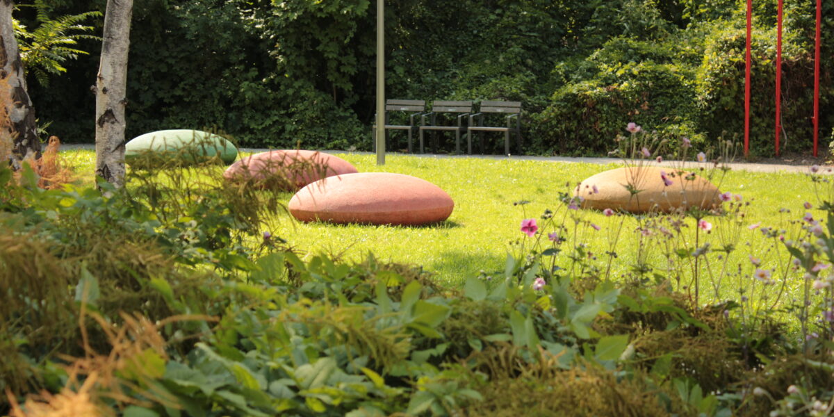 Eine grasbewachsene Parkfläche besteht aus drei großen ovalen Steinen, zwei Bänken im Hintergrund und einigen Blumen, Bäumen und roten Pfosten auf der rechten Seite.