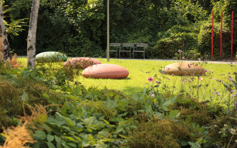 Eine grasbewachsene Parkfläche besteht aus drei großen ovalen Steinen, zwei Bänken im Hintergrund und einigen Blumen, Bäumen und roten Pfosten auf der rechten Seite.