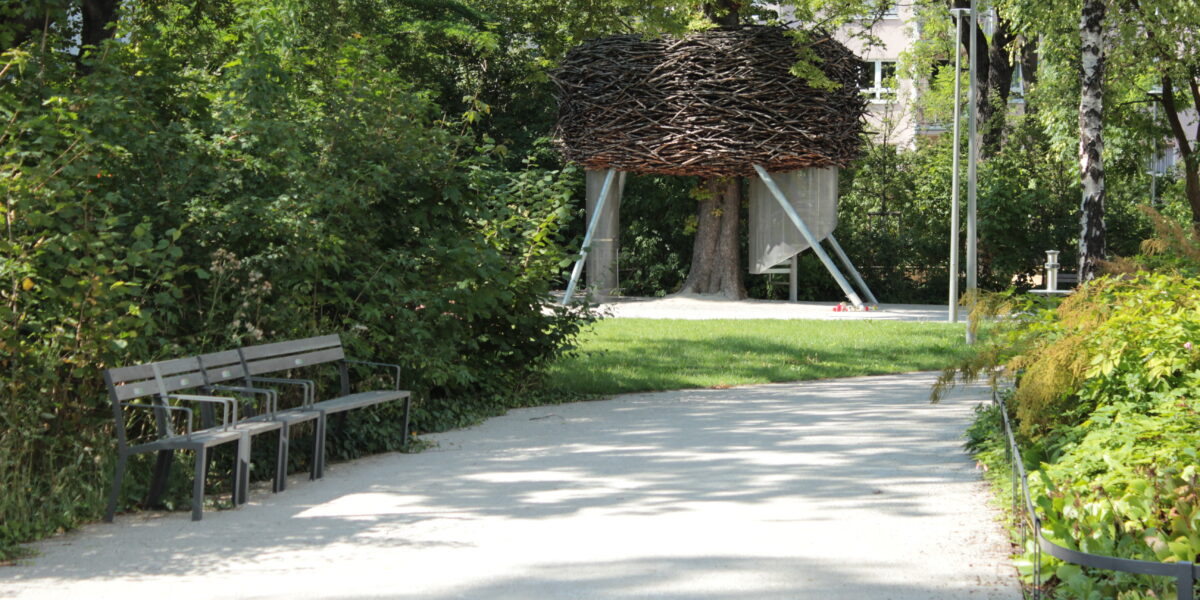 Ein Kiesweg in einem Park führt zu einer großen, erhöhten, nestartigen Holzkonstruktion mit Metallbeinen, umgeben von grünen Bäumen und Büschen. Auf der linken Seite des Weges stehen Bänke.