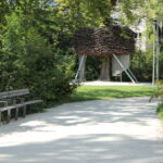 Ein Kiesweg in einem Park führt zu einer großen, erhöhten, nestartigen Holzkonstruktion mit Metallbeinen, umgeben von grünen Bäumen und Büschen. Auf der linken Seite des Weges stehen Bänke.