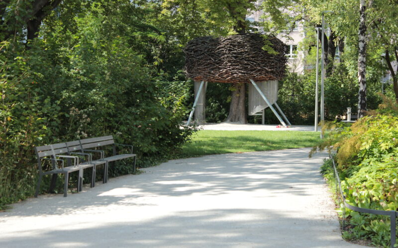 Ein Kiesweg in einem Park führt zu einer großen, erhöhten, nestartigen Holzkonstruktion mit Metallbeinen, umgeben von grünen Bäumen und Büschen. Auf der linken Seite des Weges stehen Bänke.