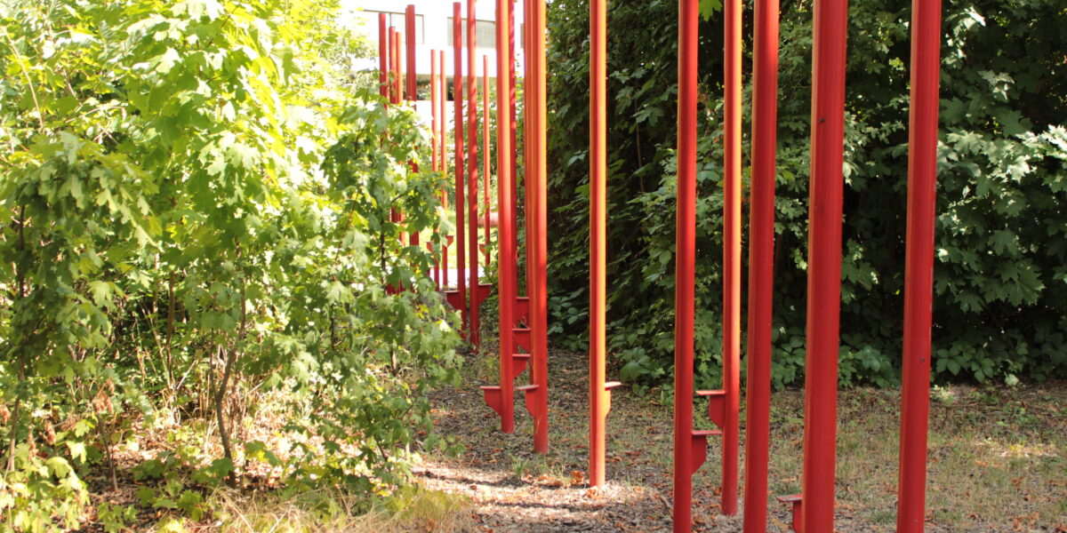 Eine Reihe hoher roter Metallstangen führt durch einen schattigen Außenbereich, der von dichtem grünen Laub und trockenen Blättern auf dem Boden umgeben ist.