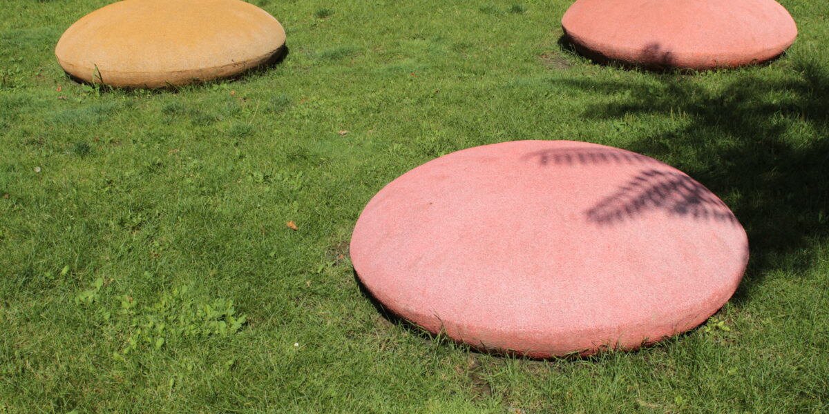 Drei große, abgerundete Betonformen - zwei rosafarbene und eine gelbe - liegen in einer Parkanlage auf dem Rasen, wobei die nächstgelegene Form teilweise von einem Schatten verdeckt wird.